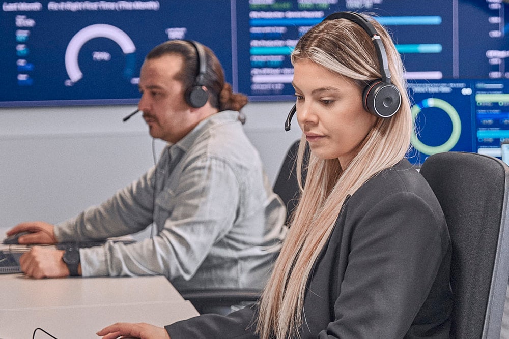  Două persoane cu căști lucrează la computere într-o sală de control modernă.