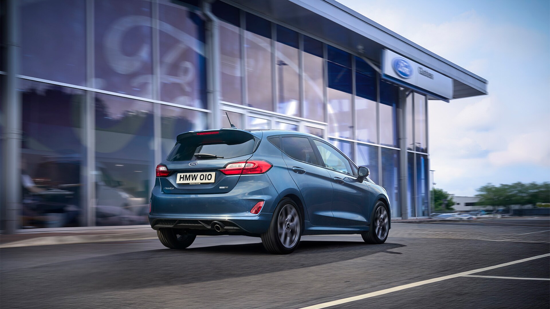 Blue hatchback car driving past a Ford dealership.