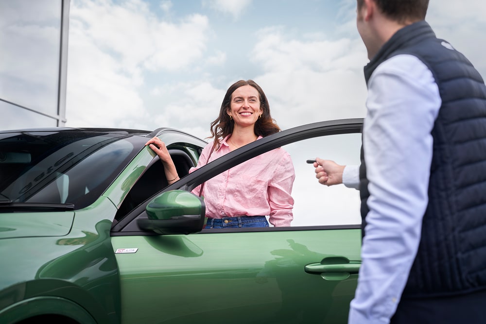 O femeie într-o cămașă roz care zâmbește lângă o mașină verde, cu un reprezentant Ford care îi înmânează cheile de la mașină.
