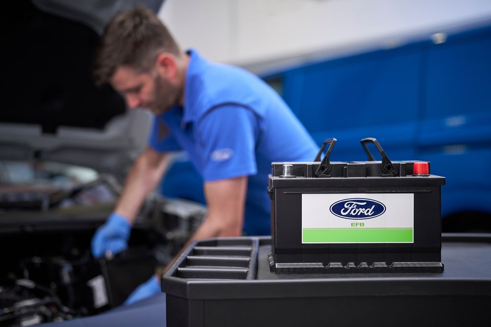 Un mecanic Ford care lucrează la un motor de mașină, cu o baterie Ford în prim-plan.