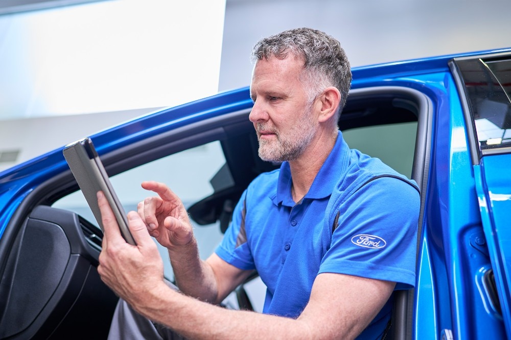 Un tehnician Ford stă într-un vehicul Ford albastru, folosind o tabletă pentru a verifica sistemele vehiculului.