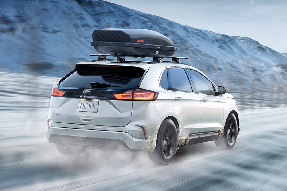 Ford Edge alb cu portbagaj de plafon, conducând pe un drum montan înzăpezit, cu efect de mișcare.