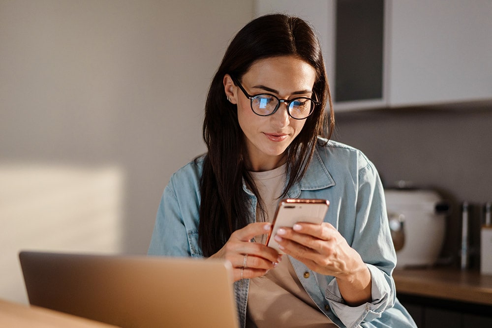 Persoană cu ochelari care privește un smartphone, cu un laptop pe o masă de lemn.
