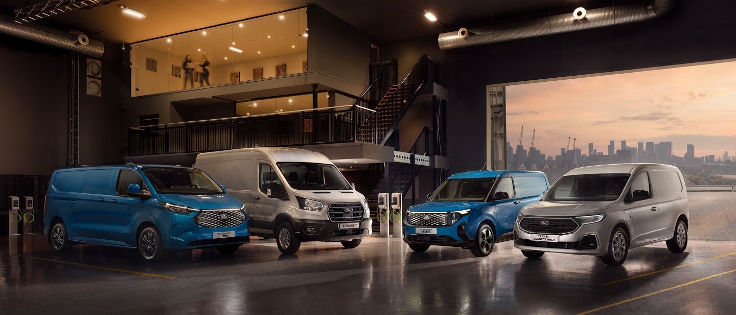 A lineup of Ford commercial vans parked inside a spacious garage, with a cityscape visible through the open doorway.