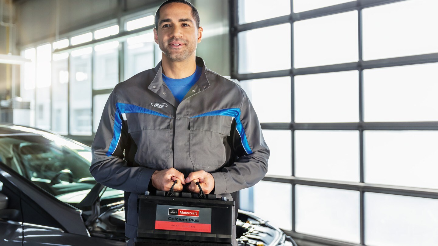 Un mecanic în uniformă Ford ținând o baterie auto, stând în fața unei mașini cu capota deschisă într-un service auto.