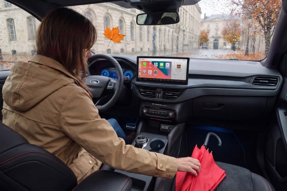 O femeie în jachetă bej întinde mâna spre o umbrelă roșie într-un Ford Kuga, într-o zi ploioasă de toamnă.
